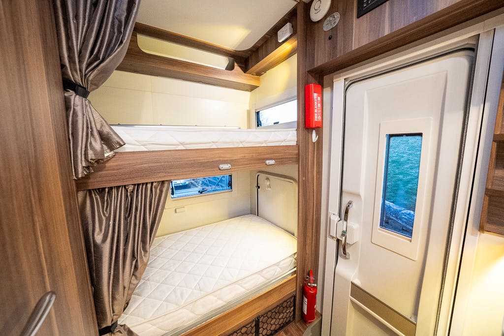 The image shows the interior of a 2016 Roller Team Auto-Roller 707 Low Line camper van featuring a bunk bed setup with two mattresses. There are windows with curtains near each bunk, a door with a window on the right, and a fire extinguisher mounted next to the door.
