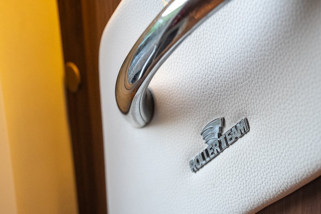 Close-up of a metallic handle attached to a white, textured surface, with "ROLLER TEAM" and a logo embossed below it. The background features a wooden section and a small circular switch or button, typical of the 2016 Roller Team Auto-Roller 707 Low Line.