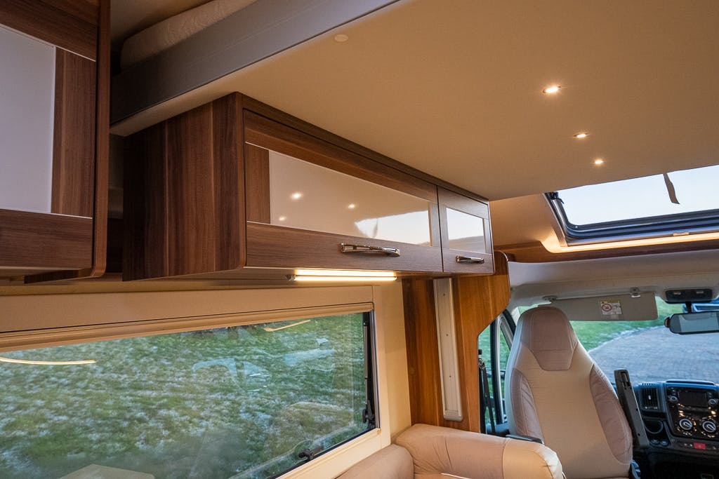 Interior view of a 2016 Roller Team Auto-Roller 707 Low Line motorhome showcasing wooden overhead cabinets with white paneling, LED lighting strips beneath, a panoramic window with a blind partially drawn, and a driver's seat with a headrest. The overall aesthetic is modern and clean.