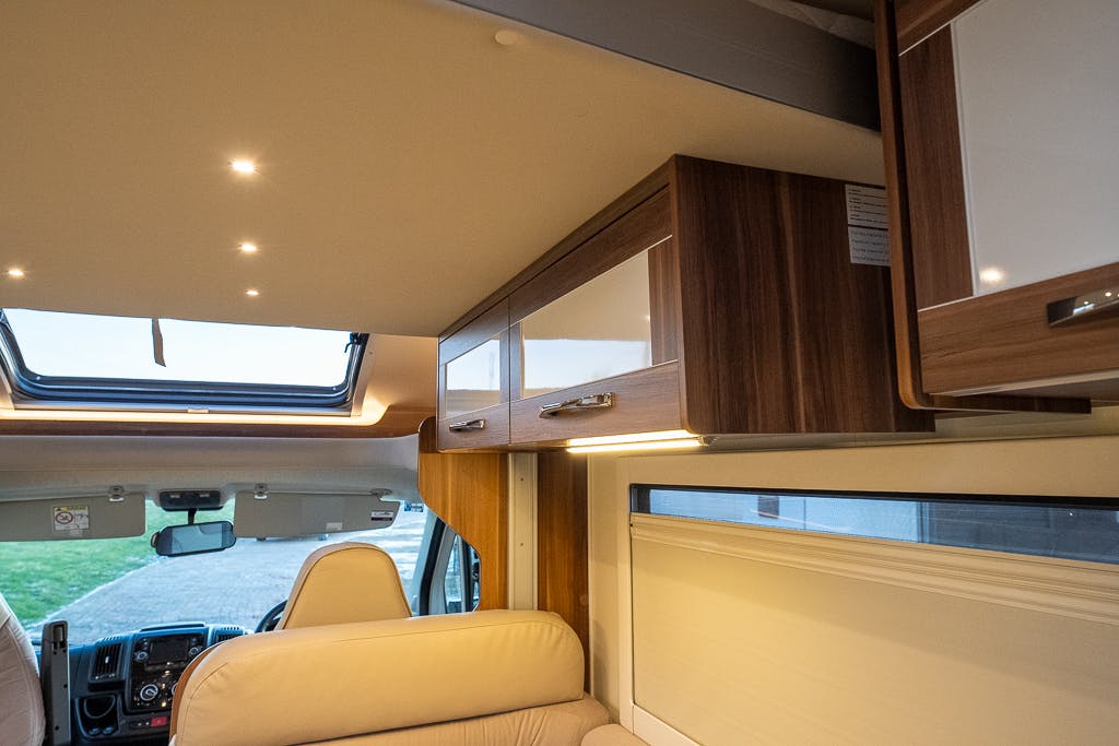 Interior view of a 2016 Roller Team Auto-Roller 707 Low Line RV showcasing the driver's area, overhead wooden cabinets with a glossy finish, and a seating area with beige upholstery. The ceiling boasts recessed lighting, and a large window is partially visible above the seating.