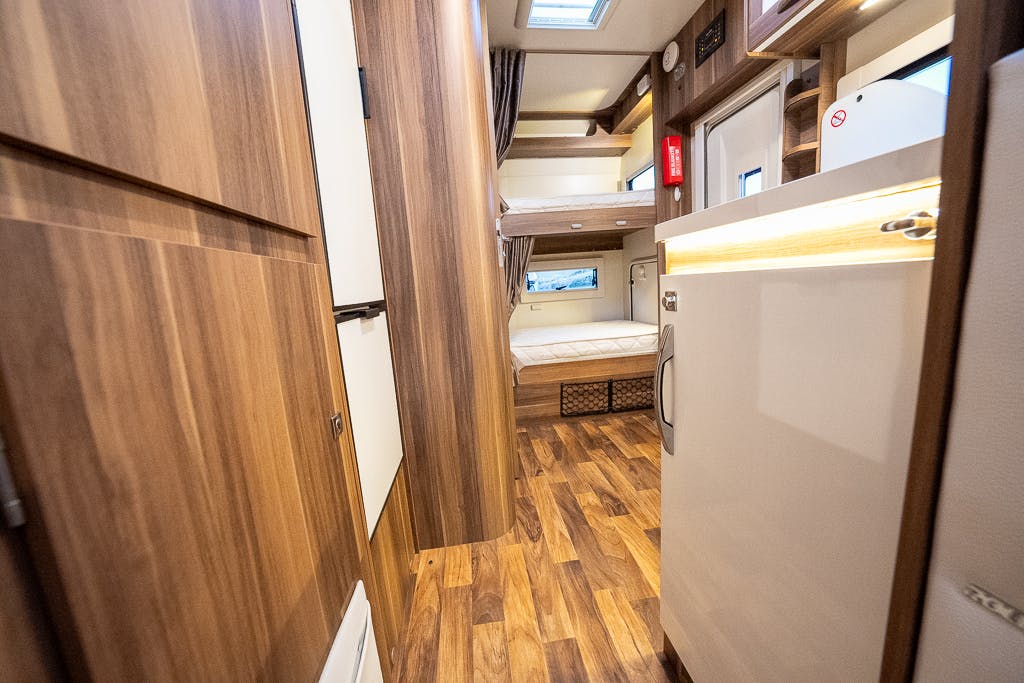 Interior view of the 2016 Roller Team Auto-Roller 707 Low Line featuring wood paneling and flooring. A small kitchen area with a sink and refrigerator is on the right, leading to a living space with a bunk bed and window. The design is modern with ample light, creating an inviting atmosphere.
