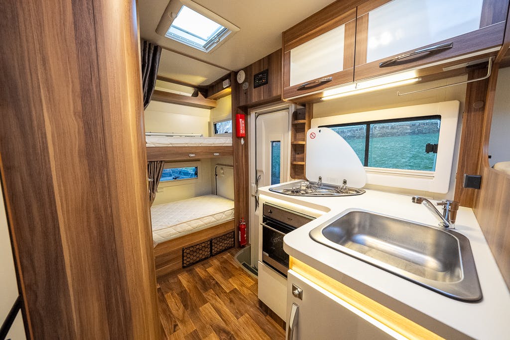 The interior of the 2016 Roller Team Auto-Roller 707 Low Line camper van features a small kitchen area with a sink, stove, and oven on the right. Opposite the kitchen is a window. At the back, there are two bunk beds. The floor is made of wood, and a fire extinguisher is mounted near the beds.