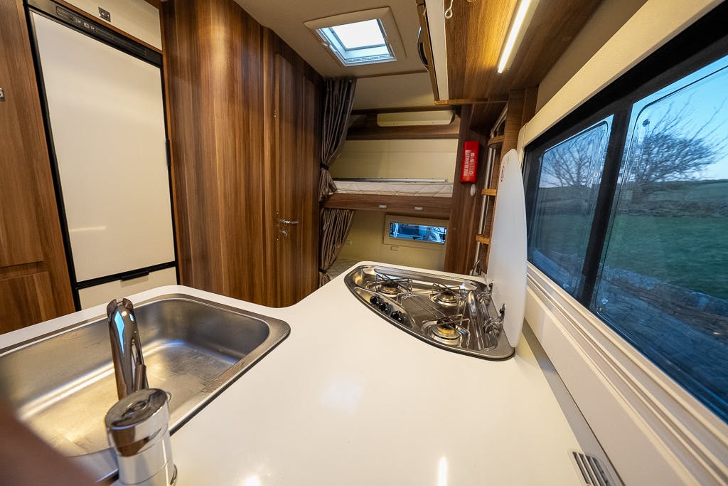 The interior of the 2016 Roller Team Auto-Roller 707 Low Line caravan shows a kitchen area with a sink, gas stove, and white countertop. A fridge is on the left, with a bunk bed visible in the background. Wooden cabinets and a window with a view of trees are also present.