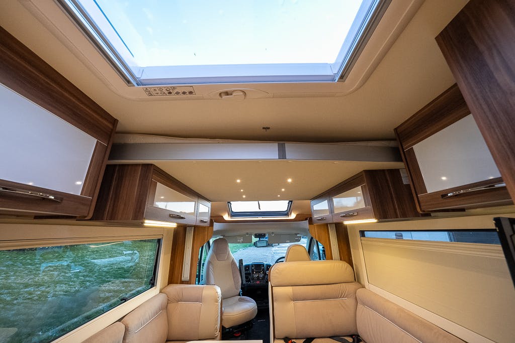 The interior of the 2016 Roller Team Auto-Roller 707 Low Line motorhome features beige leather seating, a skylight, and several wooden cabinets. The driver's area is visible at the front, and the side windows are open, offering a scenic view of the outside grassy area.