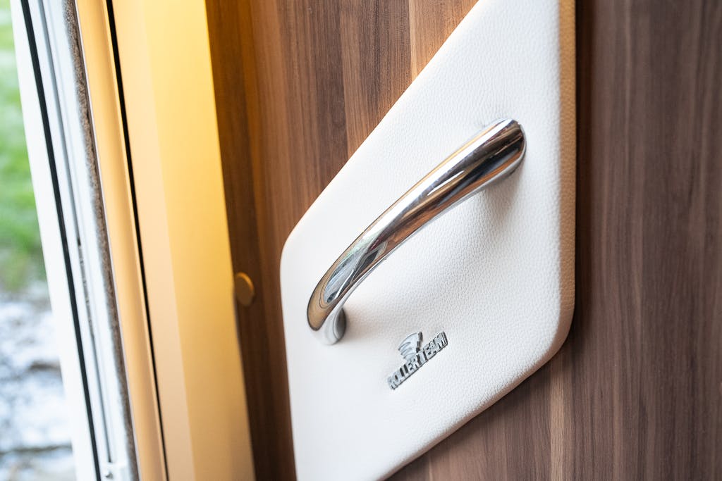 Close-up of a polished metal handle on a white surface, mounted on a wooden panel in the 2016 Roller Team Auto-Roller 707 Low Line. The surface has an oval white base with the text "MALIBU" visible. There is a window with a view of greenery and gravel on the left.
