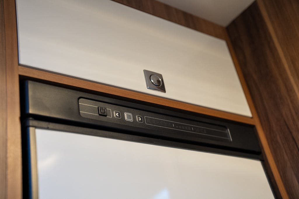 This image shows the upper section of a refrigerator or freezer integrated into wooden cabinetry, reminiscent of the sleek design found in a 2016 Roller Team Auto-Roller 707 Low Line. The appliance has a white door with a black control panel featuring various buttons and symbols, and a metallic handle centered on the door.
