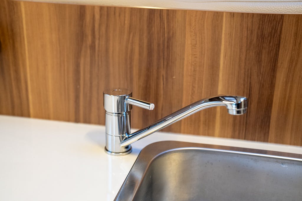 A modern, metallic kitchen faucet with a single lever handle is positioned above a stainless steel sink in the 2016 Roller Team Auto-Roller 707 Low Line. The background features wooden paneling, adding a warm contrast to the sleek design of the fixture.