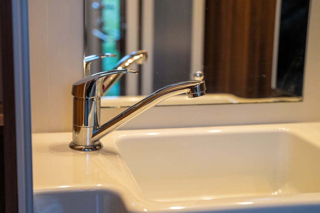 A chrome faucet over a white sink is shown in the compact, modern bathroom of the 2016 Roller Team Auto-Roller 707 Low Line. The sleek design includes a single lever for controlling water flow, perfectly reflecting in the mirror above.