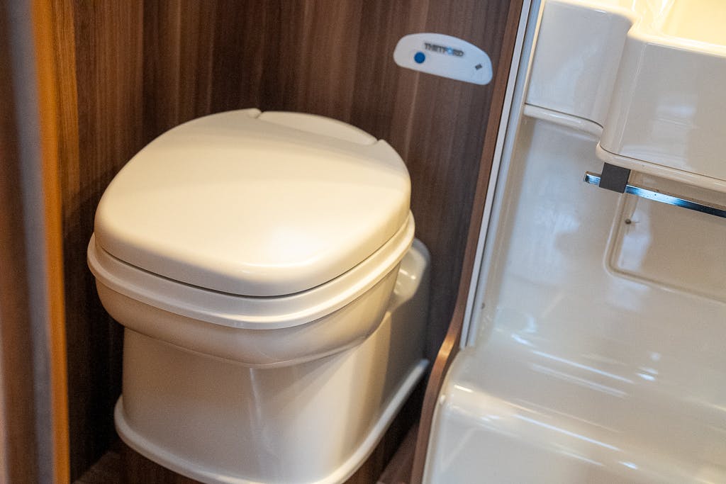 A white toilet with a closed lid is set against a wooden wall inside the 2016 Roller Team Auto-Roller 707 Low Line. Nearby, there is a white section with a door handle visible on the edge. The area appears to be a small bathroom or restroom space.