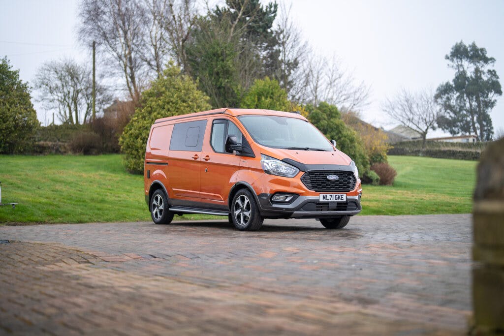 An orange 2021 Ford Transit Custom Camper is parked on a brick driveway. In the background, there is a grassy area with trees and bushes, and the sky is overcast. The van's license plate reads "ML 70 WE.