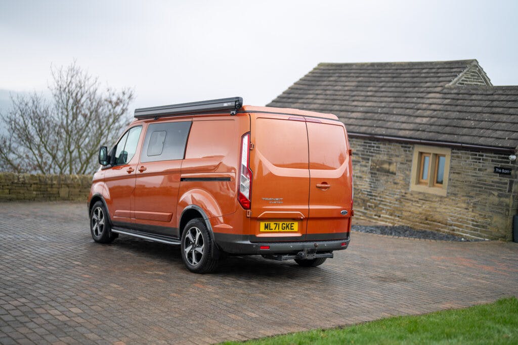 A 2021 Ford Transit Custom Camper, with an orange exterior and a black roof rack, is parked on a stone-paved driveway next to a building with a stone wall and a tiled roof. The background shows a cloudy sky and leafless trees. The van's number plate reads "ML 71 GKE.