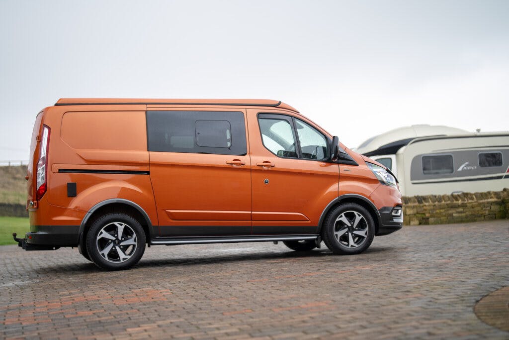 A side view of an orange 2021 Ford Transit Custom Camper parked on a paved surface. The van has black side mirrors, door handles, and wheel arches, and alloy wheels. In the background, there is a white RV and a stone wall. The sky is overcast.