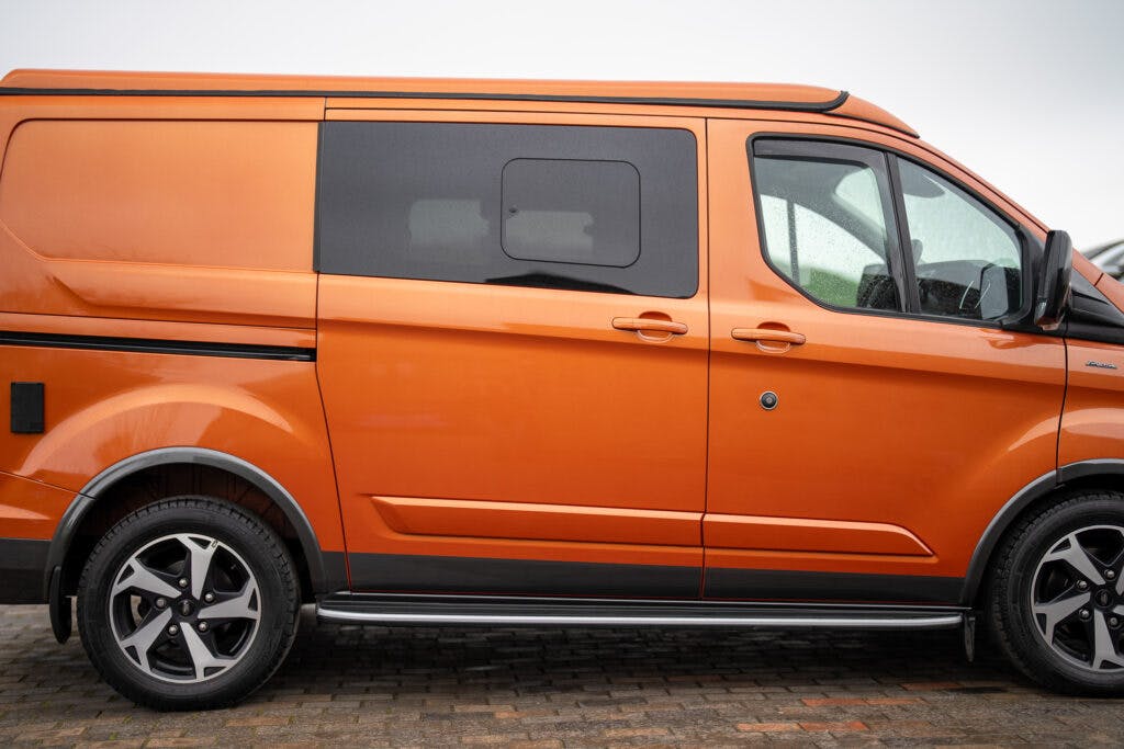 A parked orange 2021 Ford Transit Custom Camper with black-tinted windows is displayed from a side profile. The van has a modern design, alloy wheels, and a black stripe running along the sides. The ground is paved with bricks, and the background is cloudy.