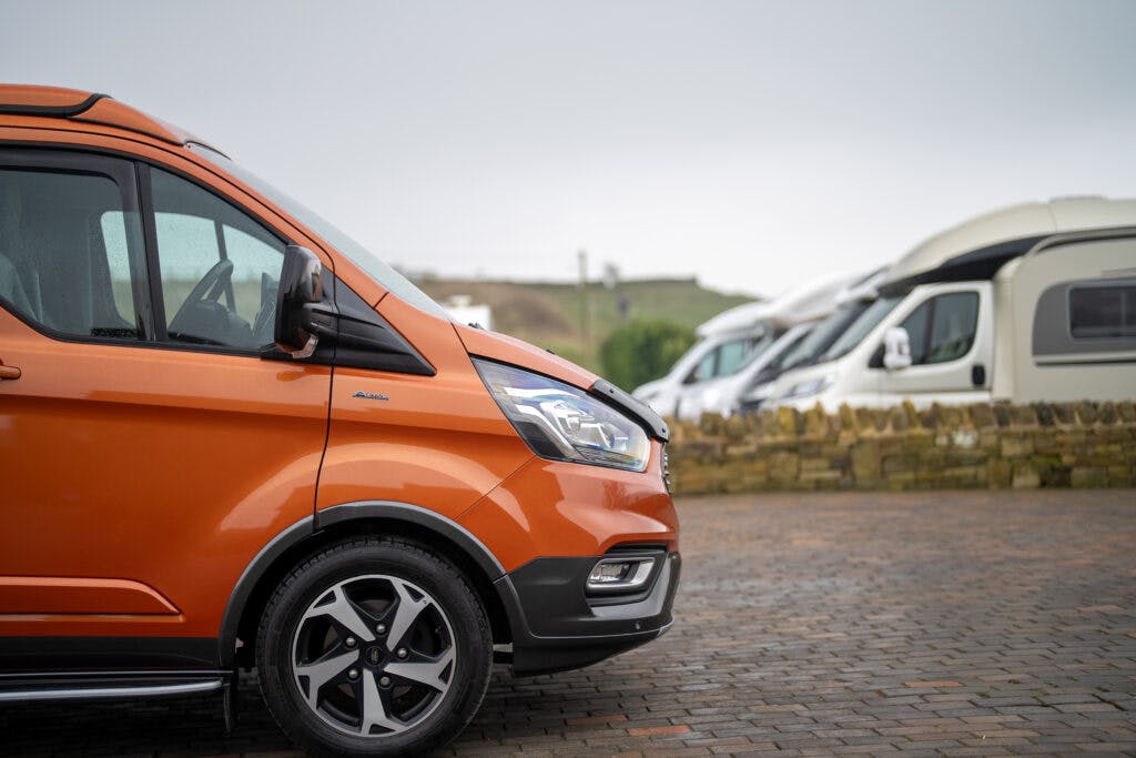A close-up of an orange 2021 Ford Transit Custom Camper parked on a cobblestone surface, with several other campervans visible in the background. The sky is overcast, and a low stone wall is seen in the distance.
