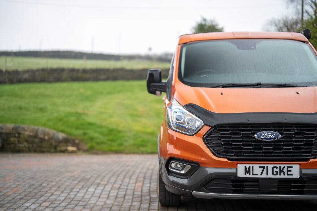 A bright orange 2021 Ford Transit Custom Camper is parked on a brick driveway. The vehicle is shown from the front, displaying its headlights, grille, and part of the license plate with the registration number "ML 71 GKE." A grassy field and stone wall are visible in the background.