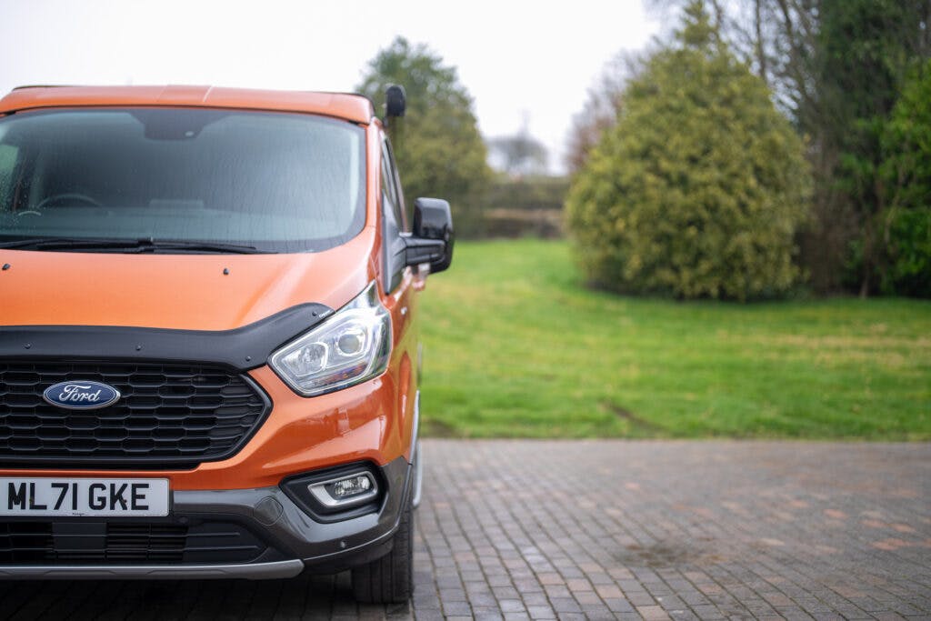 A partially visible 2021 Ford Transit Custom Camper, painted in a vibrant orange, is parked on a stone driveway with a grassy yard and lush trees in the background. The license plate reads "ML71 GKE." The scene appears to be outdoors during a cloudy day.
