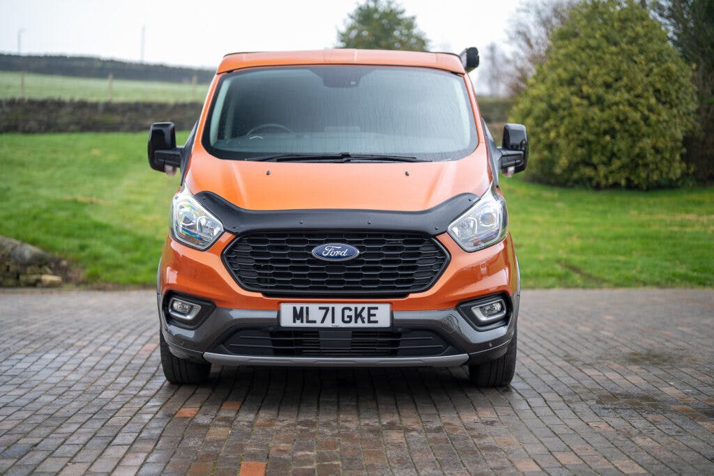 A front view of a 2021 Ford Transit Custom Camper, painted in vibrant orange, parked on a brick driveway. The vehicle's headlights and front grille are visible, along with the license plate reading "ML 71 GKE." The background showcases a grassy area with trees and a stone wall.