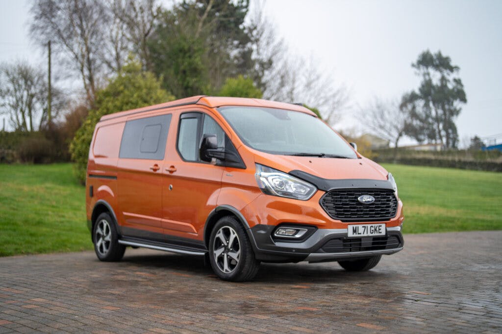 An orange 2021 Ford Transit Custom Camper van is parked on a paved surface in an outdoor setting. The vehicle has a UK license plate reading "ML71 GKE." In the background, there are trees, bushes, and a grassy area under a cloudy sky.