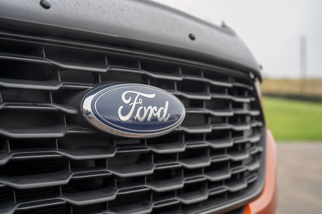 Close-up view of the front grille of a 2021 Ford Transit Custom Camper, featuring the blue and silver Ford logo in the center. The grille has a black honeycomb pattern, with part of an orange exterior visible around the edges. The background shows a blurred grassy field.