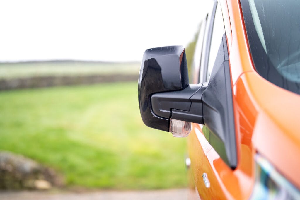 Close-up of a side mirror attached to an orange 2021 Ford Transit Custom Camper. The background features a blurry green field and a stone wall, indicating an outdoor environment. The mirror and vehicle are wet, suggesting recent rain.