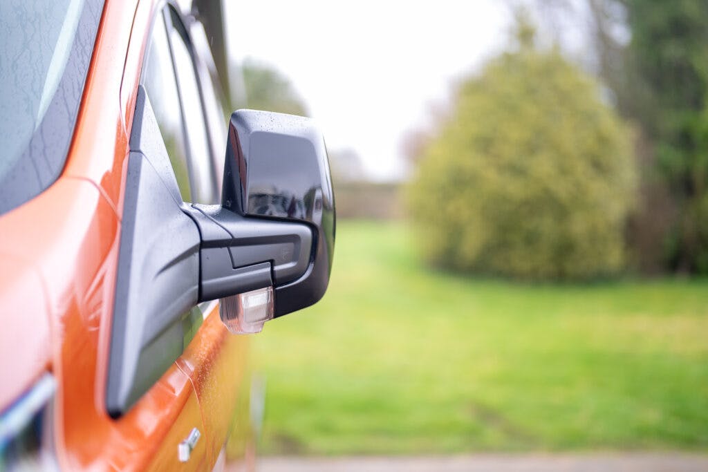 Close-up of the side mirror of a 2021 Ford Transit Custom Camper, with the background slightly out of focus. The scene appears to be outdoors with greenery and trees visible.