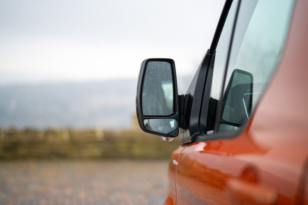 Close-up view of the side mirror of an orange 2021 Ford Transit Custom Camper with rain droplets on it. The background is slightly blurred, showing a paved surface and distant scenery.