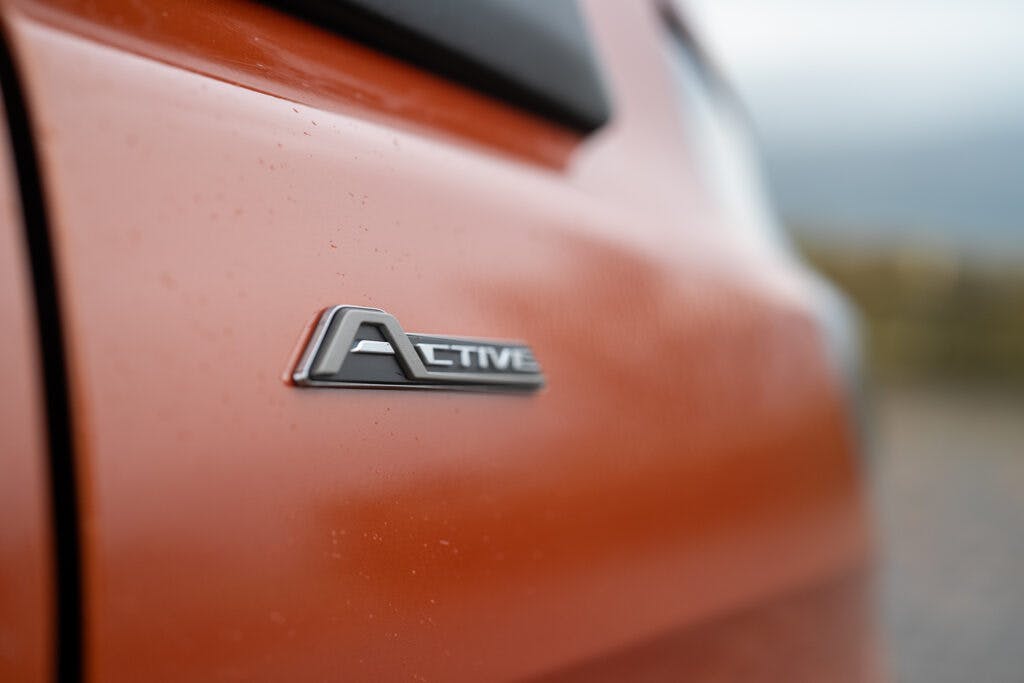 Close-up of a silver and black "Active" emblem on the side of an orange 2021 Ford Transit Custom Camper. The car's surface appears slightly wet, indicating recent exposure to moisture or rain. The background is blurred, suggesting an outdoor environment.