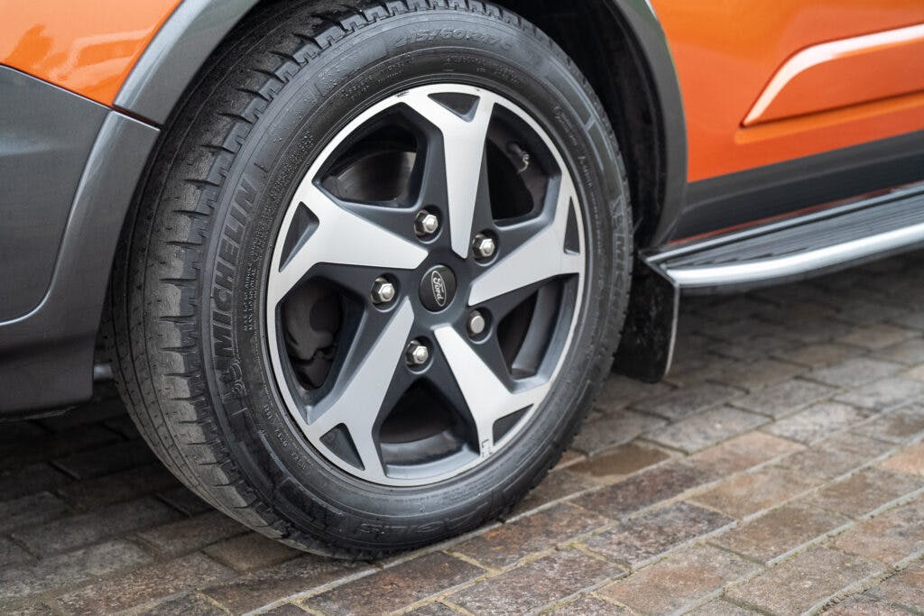 A close-up view of a 2021 Ford Transit Custom Camper's front wheel with a five-spoke alloy rim and a Goodyear tire. The camper body showcases bright orange paint, and the vehicle is parked on a cobblestone surface.