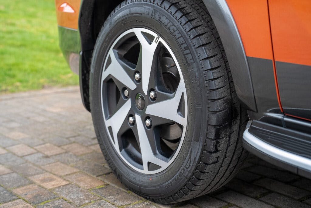 Close-up of a 2021 Ford Transit Custom Camper's rear tire mounted on an alloy wheel. The Michelin-branded tire features a five-spoke design. The camper is parked on a paved brick surface with grass visible in the background. The vehicle's body is orange.