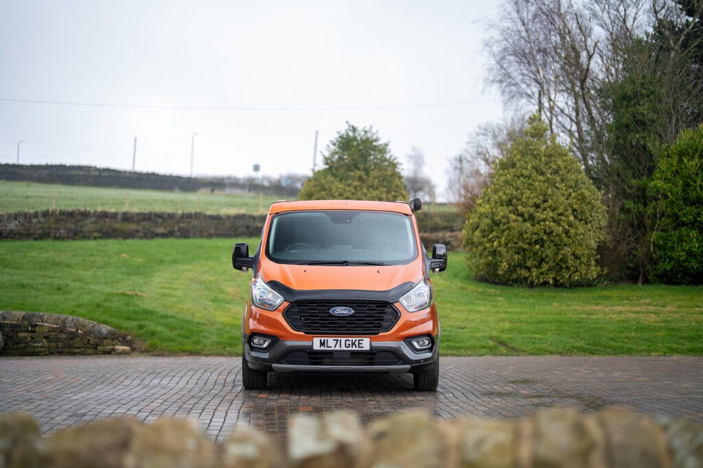 A bright orange 2021 Ford Transit Custom Camper is parked on a paved driveway. The van, boasting a UK license plate reading ML71 GKE, stands amidst green grass, stone walls, and trees under a cloudy sky.