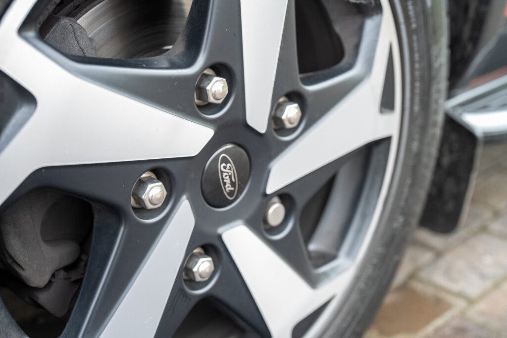 Close-up of a tire and wheel on the 2021 Ford Transit Custom Camper. The wheel features a sleek black and silver alloy design with five spokes, highlighting the "Ford" logo at its center. The tire tread and part of the car brake disc are visible, resting on a brick-paved ground.