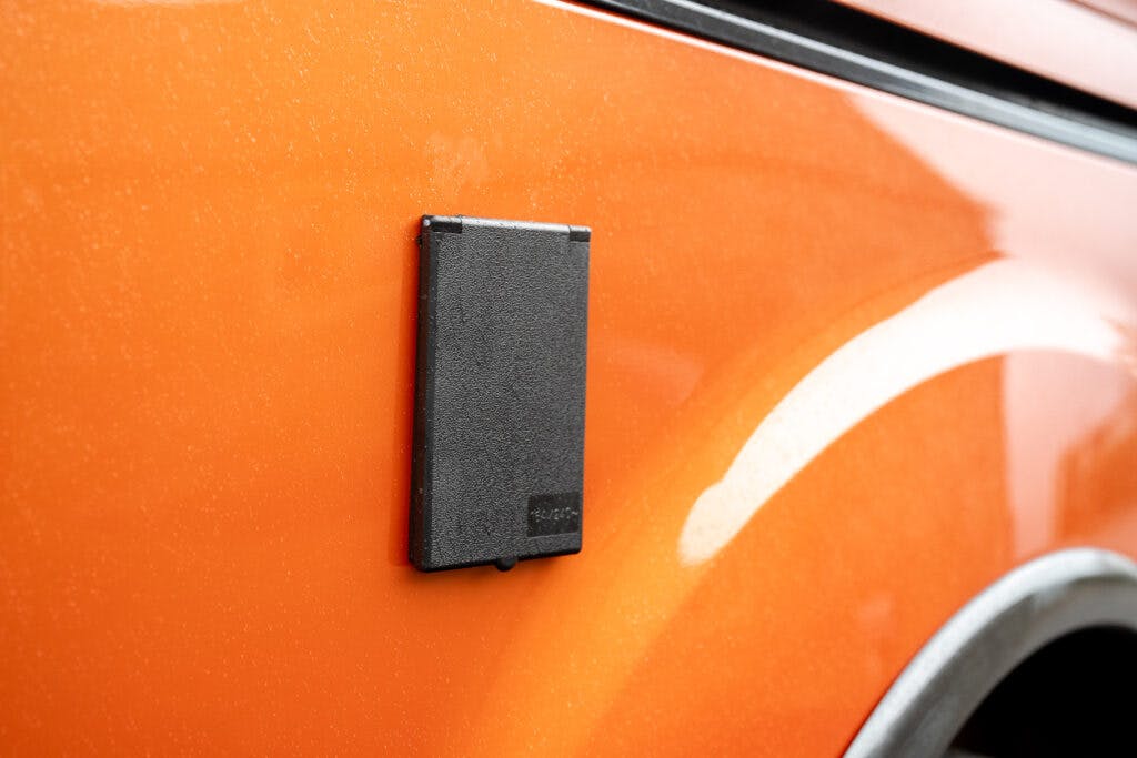 A close-up view of a 2021 Ford Transit Custom Camper's orange side panel, featuring a black rectangular fixture attached to the surface. The edge of a wheel arch is visible at the bottom right corner.