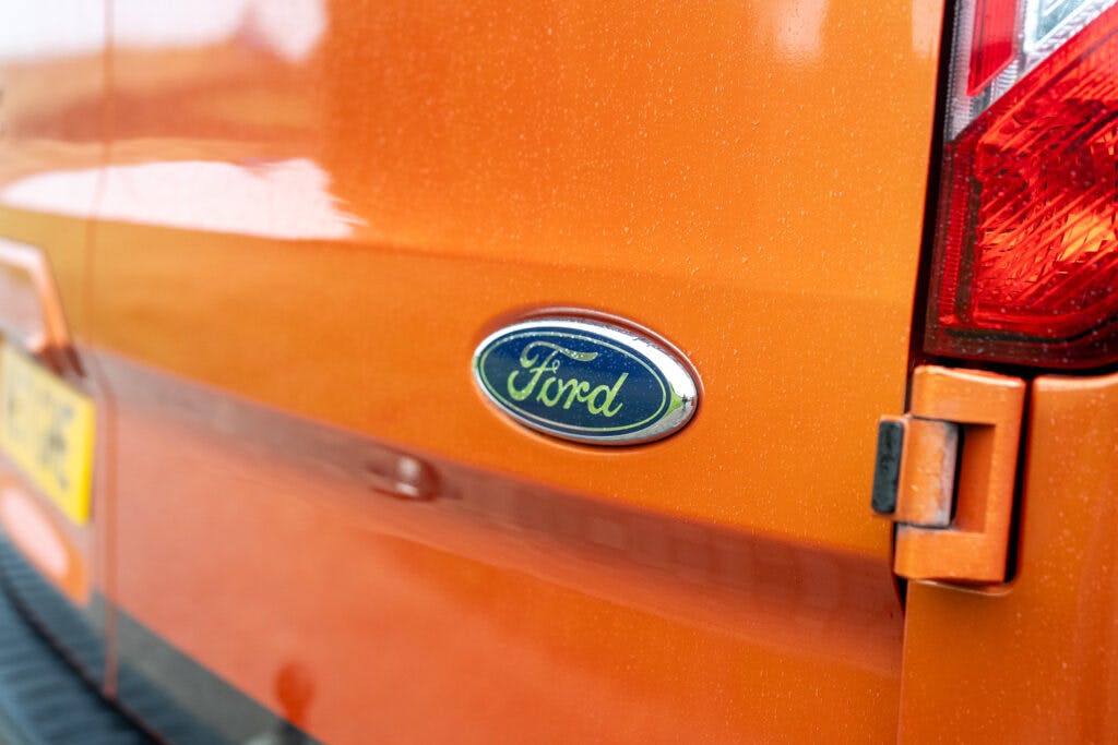 Close-up of the rear side of an orange 2021 Ford Transit Custom Camper with a Ford logo in focus. Part of a taillight and hinge are visible on the right side of the image. The vehicle appears slightly dusty or dirty. A partial view of a license plate is seen on the left.