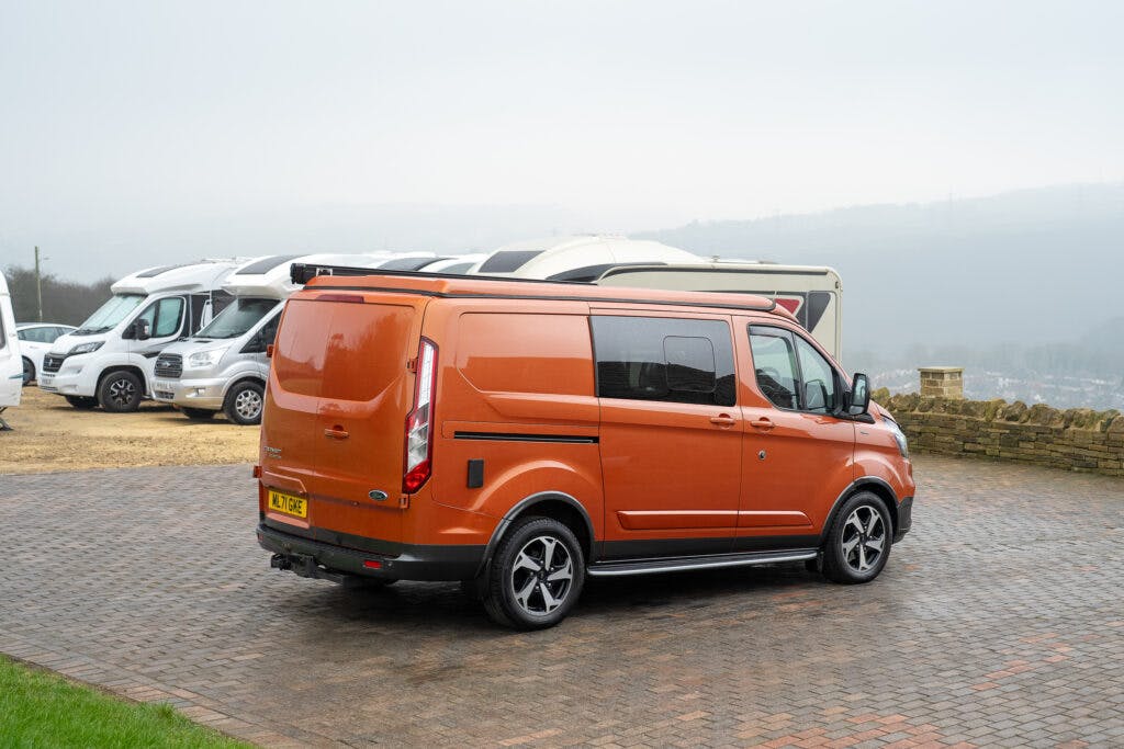 An orange 2021 Ford Transit Custom Camper is parked on a paved driveway with several white motorhomes in the background. The van is facing away from the camera, showcasing its rear and side. The sky appears overcast.