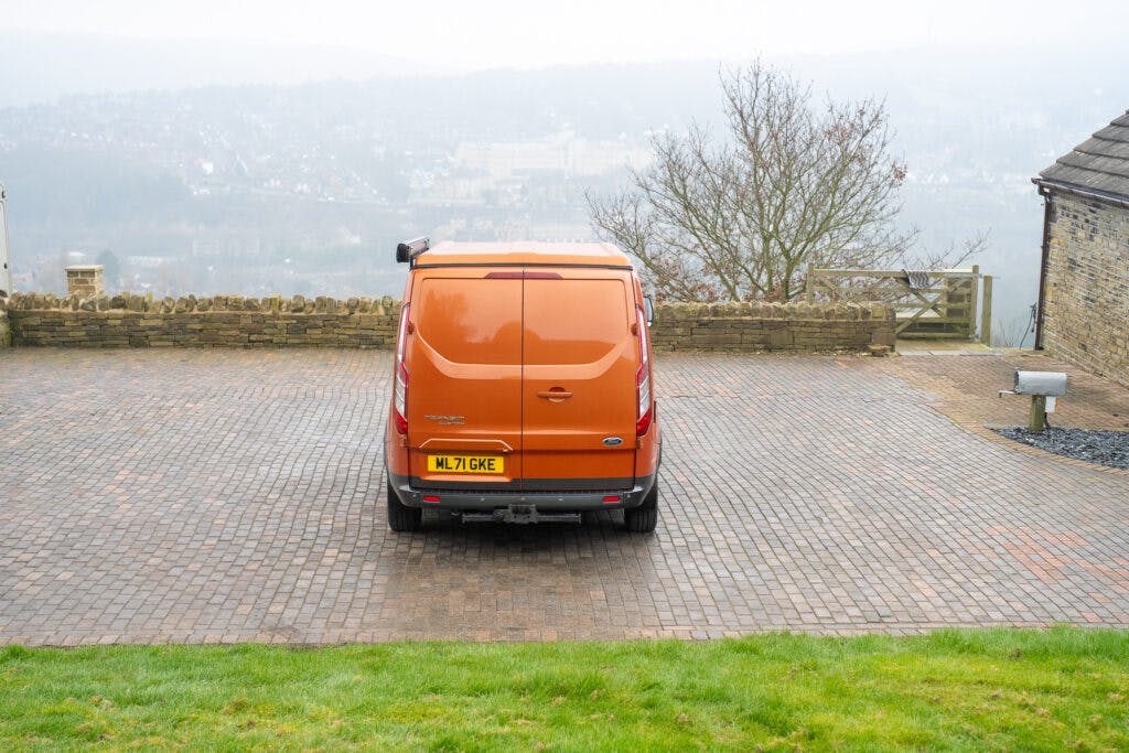 An orange 2021 Ford Transit Custom Camper with the license plate ML71 GWK is parked on a paved area next to a brick wall and a tree. The background is foggy, obscuring the view of the town or city below. A stone building is partially visible on the right.