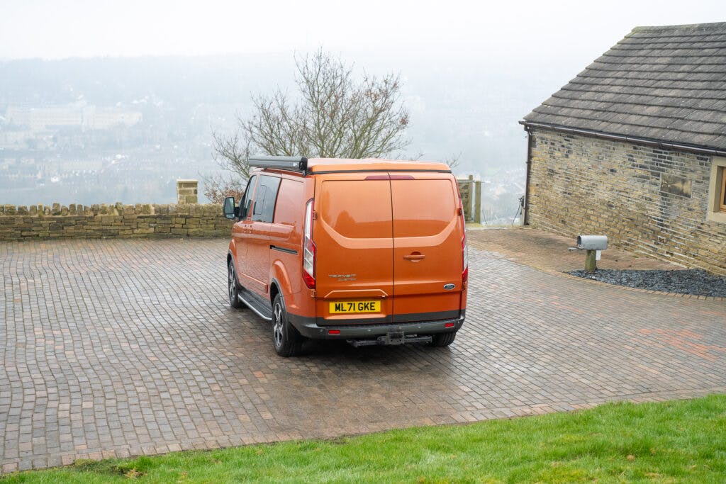 A bright orange 2021 Ford Transit Custom Camper with a license plate "ML71 GVK" is parked on a brick-paved driveway. The background includes a stone building and a partially obscured view of a foggy landscape.