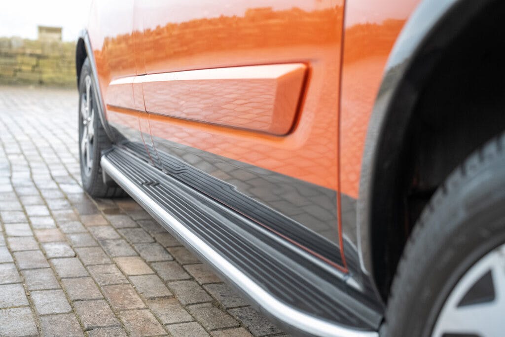 A close-up view of the side of an orange 2021 Ford Transit Custom Camper, focusing on the bottom half of the door and the side skirt. The vehicle is parked on a cobblestone driveway, with the tire and part of the wheel well visible on the right side of the image.