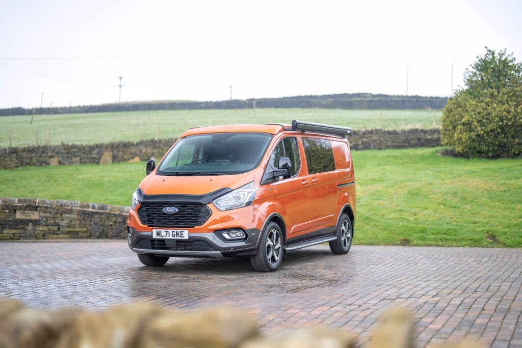 A bright orange 2021 Ford Transit Custom Camper is parked on a broad brick driveway. In the background, there is green grass, a stone wall, and a cloudy sky. The van has a registration plate with "ML71 GME" and is equipped with a side awning.