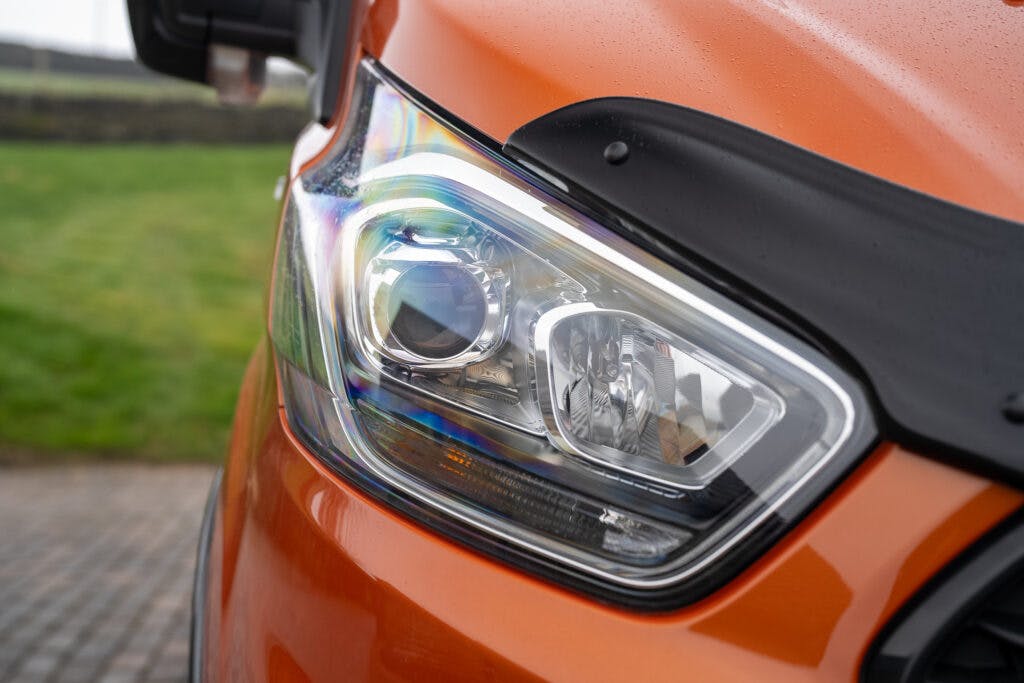 Close-up of the headlight and part of the front body of a 2021 Ford Transit Custom Camper in orange. The headlight is detailed with a clear lens and adorned with a rain protector at the top. The background includes a grassy area and a brick pavement.