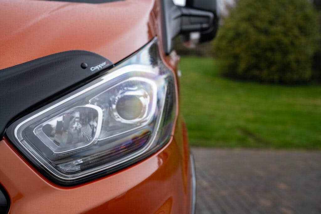 Close-up of the front left headlight of a 2021 Ford Transit Custom Camper. The headlight design features a modern, angular look with a clear lens, and the surrounding body is painted orange. The background shows a blurred grassy area and pavement.