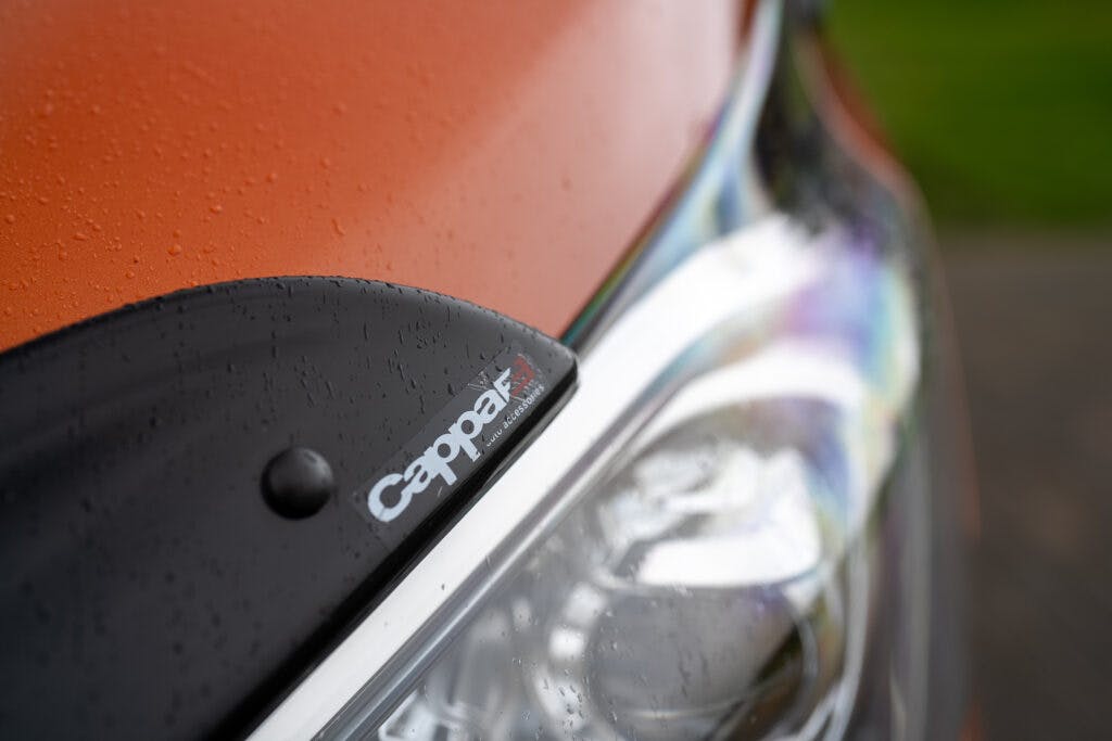 Close-up of the front part of an orange 2021 Ford Transit Custom Camper showing the headlight and a black accessory attached above it. The accessory has the brand name "Cappar" written on it. Water droplets cover the surface, suggesting recent rain.