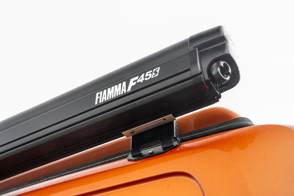 Close-up view of a Fiamma F45 awning mounted on the side of an orange 2021 Ford Transit Custom Camper. The awning is black with white branding and appears to be retracted. The background is overcast, highlighting the details of the awning and its mount.