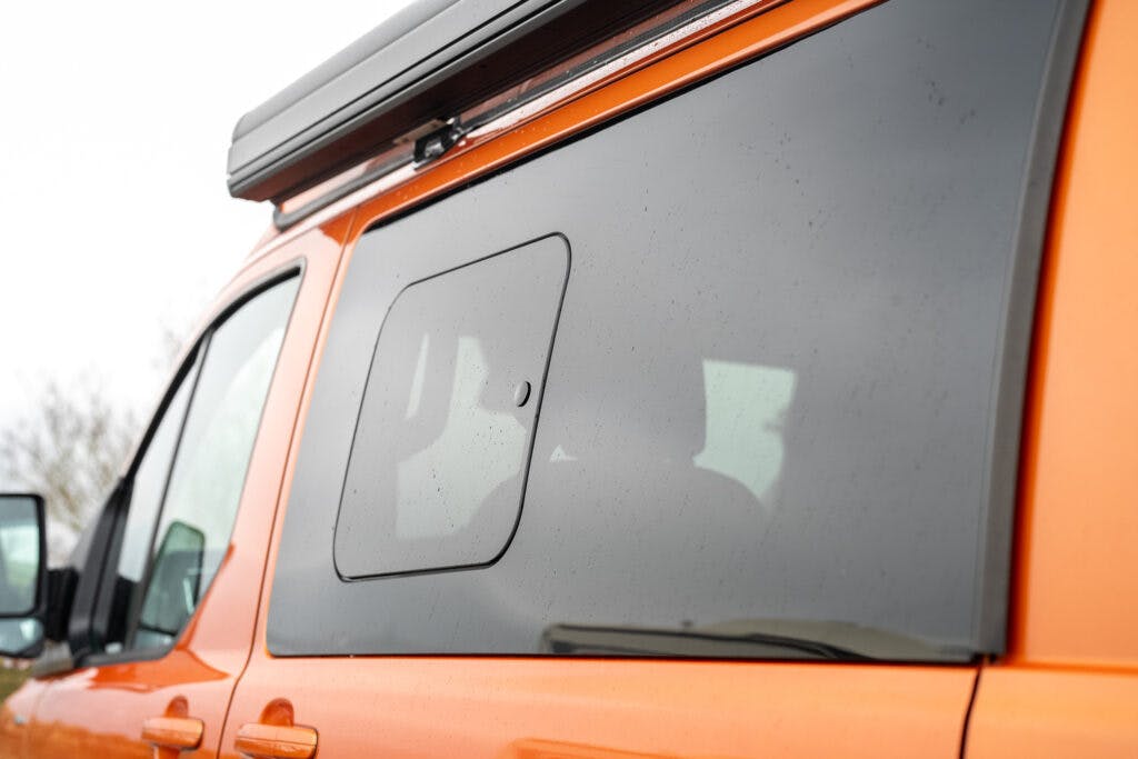 A close-up view of the side window of an orange 2021 Ford Transit Custom Camper van with a small rectangular hatch in the center. The hatch is closed, and the window is slightly tinted. There is a light overcast sky reflected on the window.