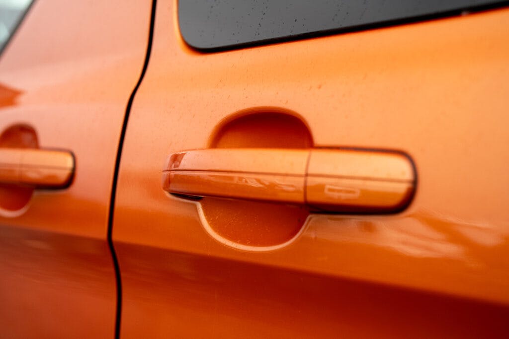Close-up of an orange car door handle on a 2021 Ford Transit Custom Camper. The handle is integrated into the sleek design of the door, which has minimal rain droplets on its surface. The camper's bright color stands out prominently.