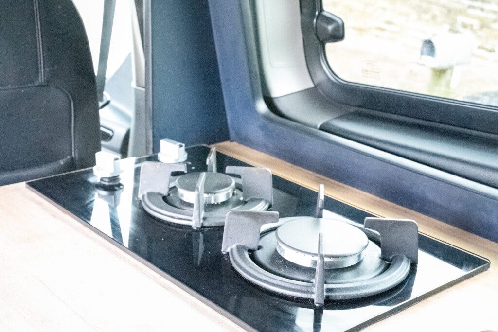 A compact kitchen area in the 2021 Ford Transit Custom Camper features a two-burner gas stove set into a wooden countertop. The stove, with black grates and control knobs, is situated next to a window offering a view of the outside.