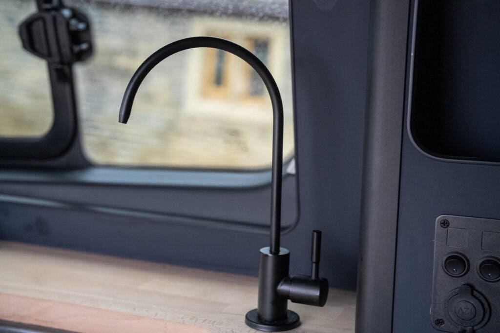 A modern, black, curved kitchen faucet is installed on a wooden countertop inside the 2021 Ford Transit Custom Camper. The background shows part of a window and a dark-colored wall with a panel featuring various buttons and switches.
