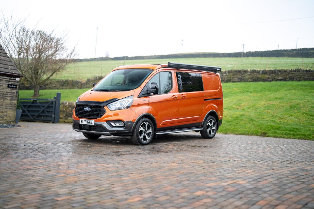 A 2021 Ford Transit Custom Camper with an orange exterior and a black roof accessory is parked on a cobblestone driveway. There is grass in the background with a fence and tree on the left side. The license plate reads "KU17 GKE." The camper van is clean and positioned facing slightly to the left.