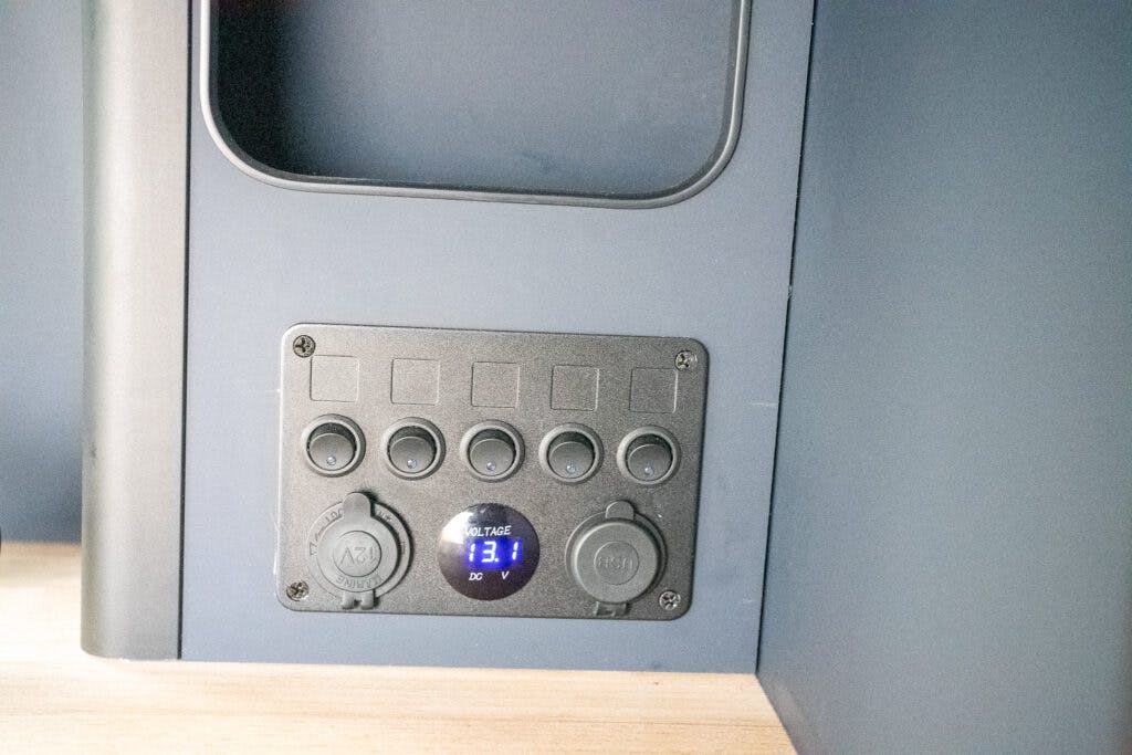 A black control panel is mounted on a blue surface inside the 2021 Ford Transit Custom Camper. The panel includes five circular buttons, two outlets labeled "A7" and "C8," and a digital display showing "13.1 DC VOLTAGE." The area surrounding the panel is partly wooden and partly blue.