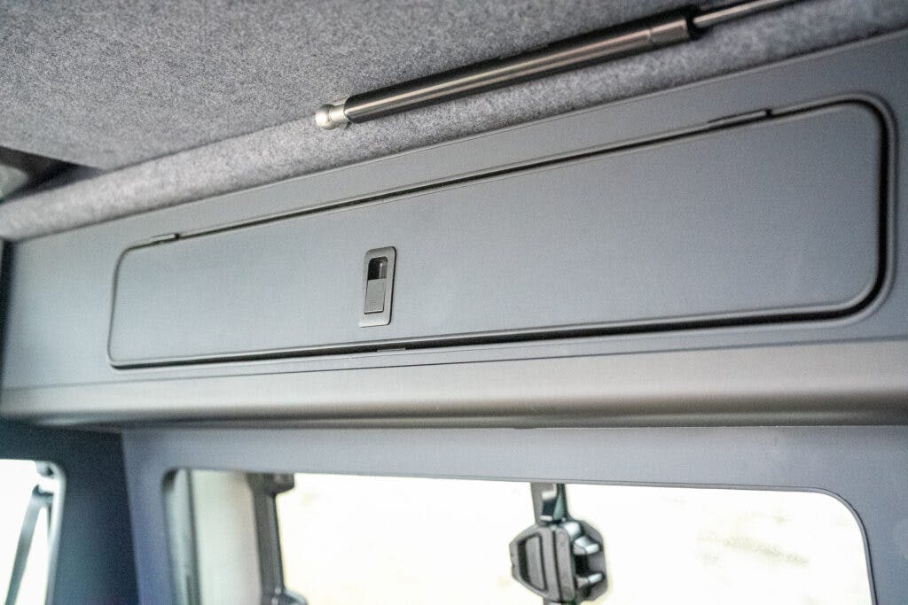 A rectangular overhead storage compartment with a lockable door is mounted inside the 2021 Ford Transit Custom Camper. Below it, a rearview mirror and part of the window are visible. The compartment appears to be made of metal or plastic, and the area is lined with gray fabric.