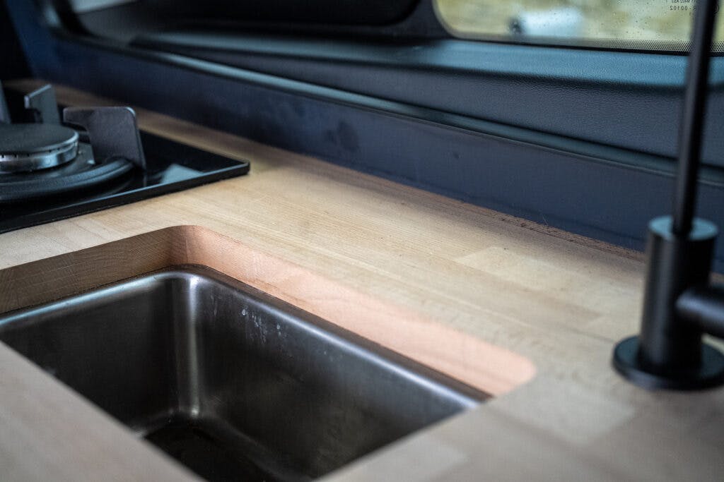 A close-up image of a wooden kitchen countertop inside a 2021 Ford Transit Custom Camper, featuring a stainless steel sink and a black gas stove in the corner. The countertop has a smooth, light finish, and there is a window in the background.